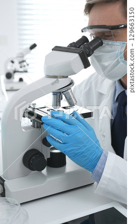 Man scientist wearing protective googles and blue gloves, is using microscope analyzing samples in modern laboratory doing scientific research. Medicine and science concept Man scientist wearing protective googles and blue gloves, is using microscope analyzing samples in modern laboratory doing scientific research. Medicine and science concept 129663150