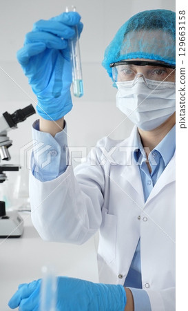 Close up of a scientist in ppe, holding a test tube with blue liquid while conducting research in a laboratory with a microscope. Medicine, health care and science concept Close up of a scientist in ppe, holding a test tube with blue liquid while conducting research in a laboratory with a microscope. Medicine, health care and science concept 129663158