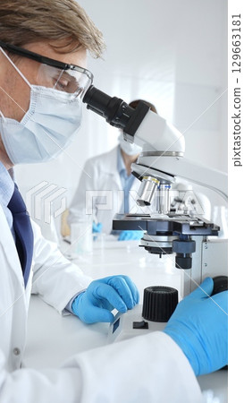 Masked male scientist analyzing sample under microscope, colleague researching nearby in sterile medical laboratory setting. Medicine, healthcare and science concept Masked male scientist analyzing sample under microscope, colleague researching nearby in sterile medical laboratory setting. Medicine, healthcare and science concept 129663181