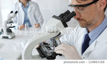 Medical scientist wearing gloves and protective glasses examining samples under a microscope in a bright laboratory filled with test tubes. Medicine, healthcare and science concept 129663188