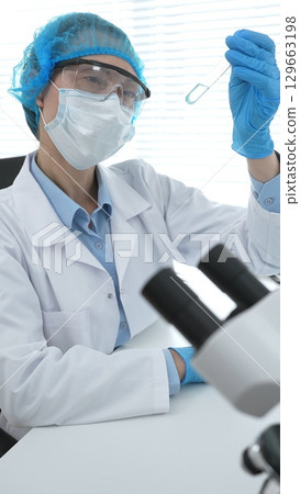 Medical science researcher wearing blue protective gear of cap, gloves and googles, is examining liquid in test tube in laboratory workspace. Medicine and health care concept Medical science researcher wearing blue protective gear of cap, gloves and googles, is examining liquid in test tube in laboratory workspace. Medicine and health care concept 129663198