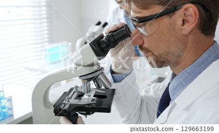 Medical scientist wearing gloves and protective glasses examining samples under a microscope in a bright laboratory filled with test tubes. Medicine, healthcare and science concept 129663199