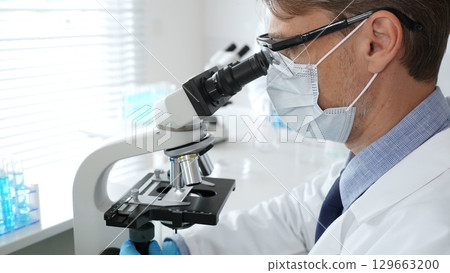 Masked scientist analyzing sample under microscope, colleague researching nearby in sterile medical laboratory setting. Medicine, healthcare and science concept Masked scientist analyzing sample under microscope, colleague researching nearby in sterile medical laboratory setting. Medicine, healthcare and science concept 129663200