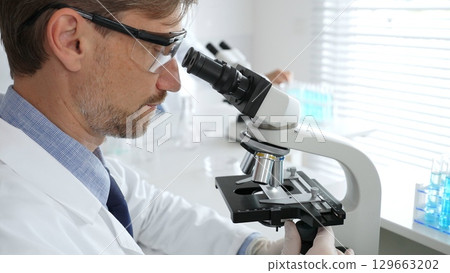 Medical scientist wearing gloves and protective glasses examining samples under a microscope in a bright laboratory filled with test tubes. Medicine, healthcare and science concept Medical scientist wearing gloves and protective glasses examining samples under a microscope in a bright laboratory filled with test tubes. Medicine, healthcare and science concept 129663202
