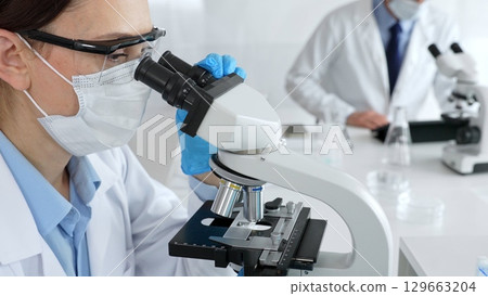 Female scientist wearing mask and gloves using microscope doing scientific research in modern laboratory with colleague working in background. Medicine, healthcare and science concept 129663204