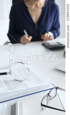 Close up of magnifying glass and calculator over financial documents opposite female accountant analyzing data, reviewing reports, binders and laptop on a white desk. Audit and taxes concept 129663205