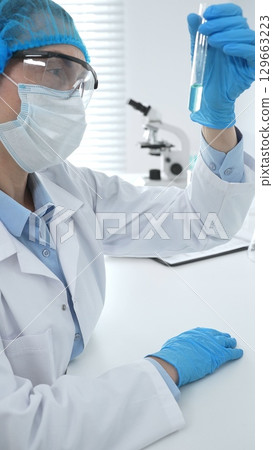 Medical science researcher wearing blue protective gear of cap, gloves and googles, is examining liquid in test tube in laboratory workspace. Medicine and health care concept 129663223