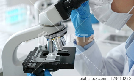 Scientific professional wearing blue protective gear examining microscope lens, performing detailed medical laboratory research. Medicine, healthcare and science concept 129663230