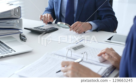 Financial analysts examining documents, using magnifying glass and calculator while studying corporate data at workplace. Audit and taxes in business 129663245