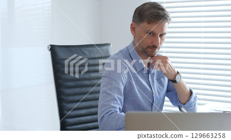 Businessman wearing blue sleek shirt is thinking and concentrating while working at the desk in a bright, modern office, demonstrating professionalism and efficiency. Business people concept Businessman wearing blue sleek shirt is thinking and concentrating while working at the desk in a bright, modern office, demonstrating professionalism and efficiency. Business people concept 129663258