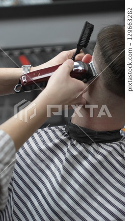 Professional barber grooming client's hair using electric clipper, delivering precise cut with skill in contemporary salon setting, close-up view Professional barber grooming client's hair using electric clipper, delivering precise cut with skill in contemporary salon setting, close-up view 129663282