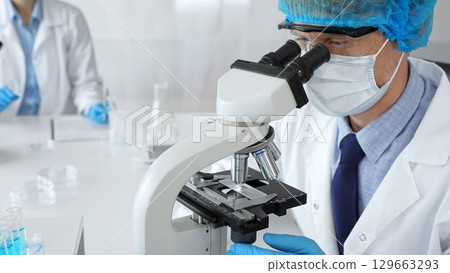 Senior male scientist wearing a protective mask, cap, and gloves, examining samples under a microscope in a modern laboratory. Medicine, healthcare and science concept 129663293
