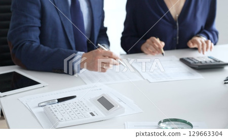 Calculator and magnifying glass over financial documents opposite financial team analyzing data, reviewing reports, collaborating at modern office workspace, close up view. Audit and taxes 129663304