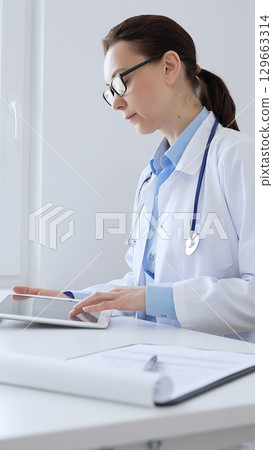 Middle aged doctor woman wearing lab coat and stethoscope using digital tablet with laptop and medical records on desk in medical office, showcasing modern healthcare technology. Medicine and health Middle aged doctor woman wearing lab coat and stethoscope using digital tablet with laptop and medical records on desk in medical office, showcasing modern healthcare technology. Medicine and health 129663314
