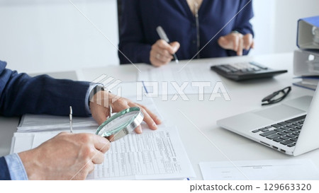 Two professional chartered accountants reviewing financial reports and analyzing data using a calculator and a magnifying glass, ensuring accuracy and compliance. Audit and taxes in business Two professional chartered accountants reviewing financial reports and analyzing data using a calculator and a magnifying glass, ensuring accuracy and compliance. Audit and taxes in business 129663320