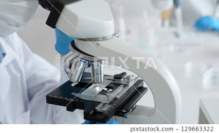 Scientific professional wearing blue protective gear examining microscope lens, performing detailed medical laboratory research. Medicine, healthcare and science concept 129663322