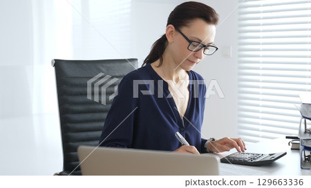 Professional female wearing black glasses, is working at desk, calculating finances, writing notes, managing business documents in contemporary workspace. Audit and taxes in business 129663336