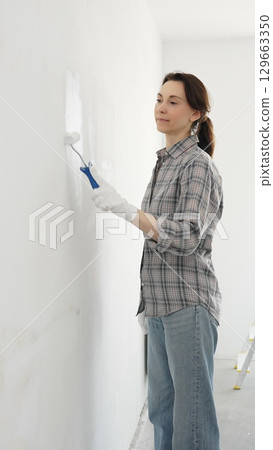 Middle aged happy woman wearing checked shirt, protective gloves, is rolling white paint onto wall, updating living space with fresh color and personal home renovation effort 129663350