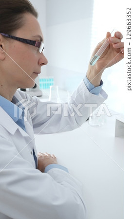 Female researcher examining clear liquid in test tube, sitting near microscopes and lab equipment within sterile scientific workspace. Medicine, healthcare and science concept 129663352