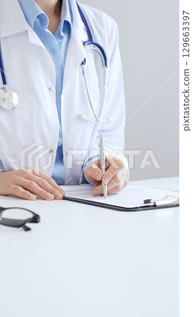 Unknown female doctor completing healthcare paperwork, clipboard positioned near laptop and stethoscope in well lit clinical workspace by window. Medicine and health care Unknown female doctor completing healthcare paperwork, clipboard positioned near laptop and stethoscope in well lit clinical workspace by window. Medicine and health care 129663397