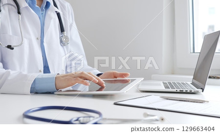 Female doctor wearing white medical coat and stethoscope using digital tablet with laptop and medical records on desk in medical office, showcasing modern healthcare technology. Medicine and helthcare 129663404