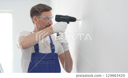 Middle aged man wearing blue overall and protective glasses, is drilling hole in white wall, wearing protective gear during home improvement project with cordless power tool 129663420