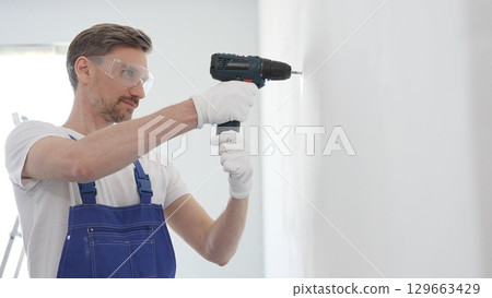 Middle aged man wearing blue overall and protective glasses, is drilling hole in white wall, wearing protective gear during home improvement project with cordless power tool 129663429