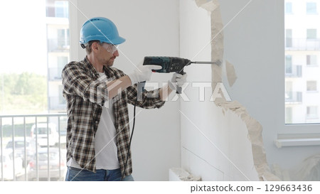 Man construction worker demolishing a wall with a hammer drill in protective gear, creating construction debris on the surrounding floor surface. Renovation concept 129663436