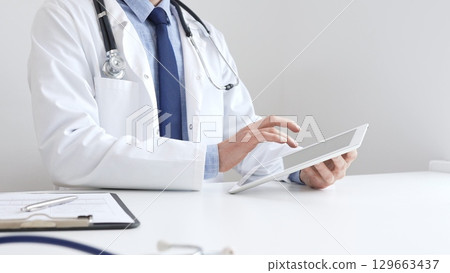 Male doctor wearing white medical coat and stethoscope is using digital tablet while sitting at desk in medical office, with medical chart and pen in the foreground. Medicine and health care concept 129663437