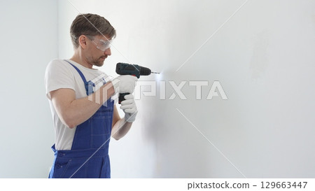 Skilled worker drilling hole in white wall, wearing protective gear during home improvement project with cordless power tool 129663447