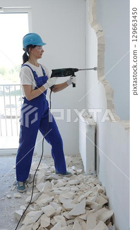 Woman construction worker wearing blue overalls, safety glasses and hardhat is demolishing interior wall with rotary hammer, creating substantial concrete debris pile during demolition process Woman construction worker wearing blue overalls, safety glasses and hardhat is demolishing interior wall with rotary hammer, creating substantial concrete debris pile during demolition process 129663450
