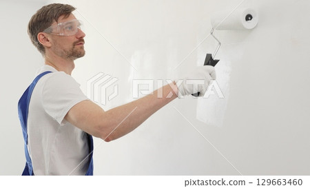Male professional painter wearing blue overall, is carefully rolling white paint on wall, creating uniform surface during interior home improvement project Male professional painter wearing blue overall, is carefully rolling white paint on wall, creating uniform surface during interior home improvement project 129663460
