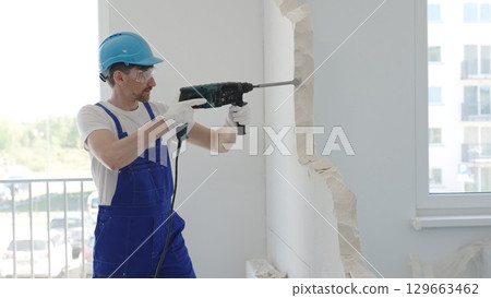 Construction worker demolishing concrete wall with rotary hammer, wearing protective safety gear, generating dust and debris during home renovation work Construction worker demolishing concrete wall with rotary hammer, wearing protective safety gear, generating dust and debris during home renovation work 129663462