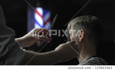 Professional barber carefully cutting hair of male customer with scissors, providing stylish and precise haircut in modern barbershop background Professional barber carefully cutting hair of male customer with scissors, providing stylish and precise haircut in modern barbershop background 129663463