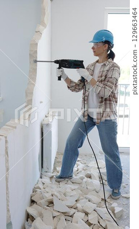 Female construction worker demolishing a wall with a hammer drill, wearing protective gear, generating rubble pile during renovation project Female construction worker demolishing a wall with a hammer drill, wearing protective gear, generating rubble pile during renovation project 129663464