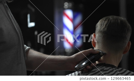 Male professional barber trimming client's hair with electric clipper, vintage illuminated barber pole visible in background, creating contemporary grooming experience 129663465
