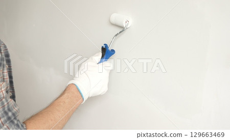 Male painter's gloved hand gripping paint roller, spreading white paint across wall during home interior transformation with professional precision, close-up view. Renovation concept Male painter's gloved hand gripping paint roller, spreading white paint across wall during home interior transformation with professional precision, close-up view. Renovation concept 129663469