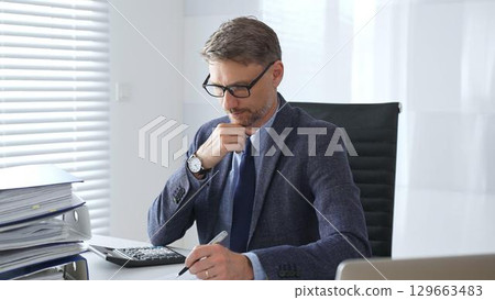 Adult businessman wearing blue suit and tail is concentrating and thinking while working at the desk in a bright, modern office, demonstrating professionalism and efficiency. Business people concept 129663483