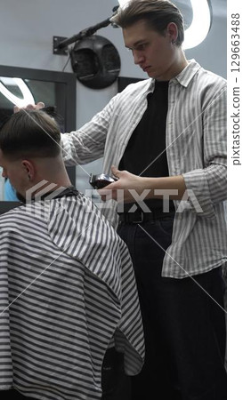 Professional barber carefully trimming customer's hair with electric clipper, providing a stylish and precise haircut in a modern barbershop environment 129663488