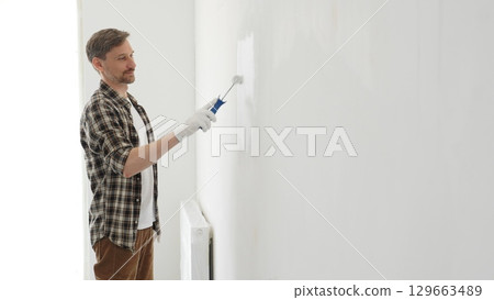 Middle aged man wearing checked shirt and protective gloves, is painting a white wall with a paint roller, working on a home renovation project, creating a fresh and clean look 129663489