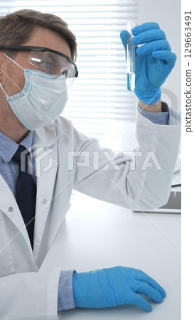 Scientist in a white lab coat, face mask, and gloves, analyzing a blue liquid in a test tube within a laboratory setting. Medicine, healthcare and science concept 129663491