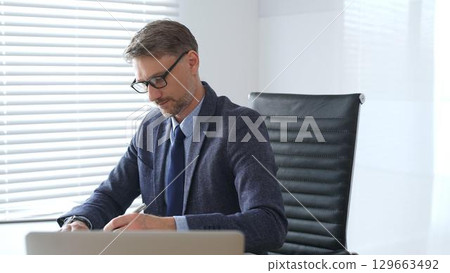 Senior businessman wearing suit, tail and black glasses is concentrating while writing on a document at his desk in a bright, modern office, demonstrating professionalism. Business people concept 129663492