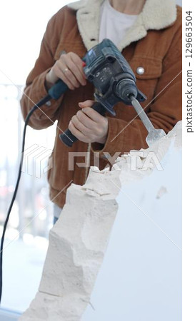 Unknown female construction worker demolishing concrete wall with rotary hammer, wearing protective safety gear. Renovation concept, closeup vertical view 129663504