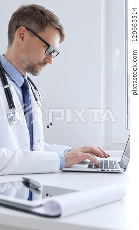 Middle aged doctor man is diligently typing on a laptop, likely updating patient records or conducting research, showcasing the intersection of healthcare and technology in medical setting. Medicine Middle aged doctor man is diligently typing on a laptop, likely updating patient records or conducting research, showcasing the intersection of healthcare and technology in medical setting. Medicine 129663514