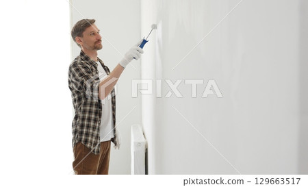 Middle aged man wearing checked shirt and protective gloves, is painting a white wall with a paint roller, working on a home renovation project, creating a fresh and clean look Middle aged man wearing checked shirt and protective gloves, is painting a white wall with a paint roller, working on a home renovation project, creating a fresh and clean look 129663517