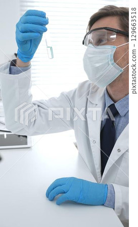 Scientist in a white lab coat, face mask, and gloves, analyzing a blue liquid in a test tube within a laboratory setting. Medicine, healthcare and science concept 129663518