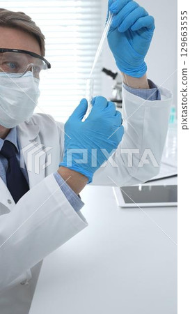 Masked male scientist in lab coat, protective glasses and blue gloves is adding reagent to test tube with microscopes and test tube rack in modern laboratory. Medicine, healthcare and science concept Masked male scientist in lab coat, protective glasses and blue gloves is adding reagent to test tube with microscopes and test tube rack in modern laboratory. Medicine, healthcare and science concept 129663555