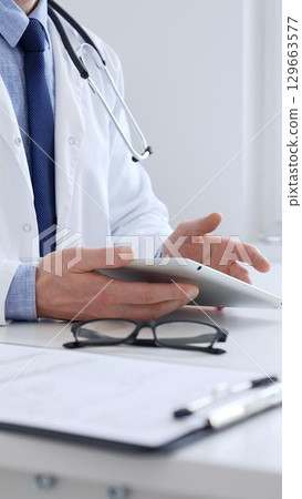 Adult male doctor wearing lab coat and stethoscope using digital tablet while sitting at desk in medical office, with medical chart and pen in the foreground, close up. Medicine 129663577