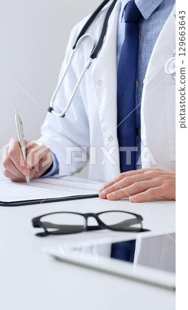 Unreckognisable doctor man writing a prescription and filling out medical records on a clipboard in an office setting. Medicine and health care concept Unreckognisable doctor man writing a prescription and filling out medical records on a clipboard in an office setting. Medicine and health care concept 129663643