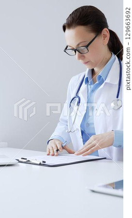 Adult female doctor completing healthcare paperwork, clipboard positioned near laptop and stethoscope in well lit clinical workspace by window. Medicine and health care Adult female doctor completing healthcare paperwork, clipboard positioned near laptop and stethoscope in well lit clinical workspace by window. Medicine and health care 129663692